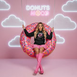 Model posing with a large pink donut inflatable against a pink wall with neon text and cloud shapes, wearing the Mood Sweet black hoodie with QR code for AR video activation.