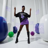 Model wearing a black t-shirt with an acai bowl design and purple skirt in a studio setting with colorful balls and white curtains.