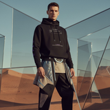 Man wearing an AR black hoodie and futuristic pants standing in a desert with geometric glass structures.