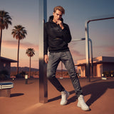 Man in black hoodie with QR code for AR video activation, and gray pants standing in a desert-like setting with palm trees and modern buildings.